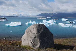 Im Vordergrund des Bildes sieht man einen Felsen, der ein bisschen wie ein lächelndes Gesicht aussieht. Im Hintergrund erstreckt sich ein weiter See mit großen, treibenden Gletscherstücken.