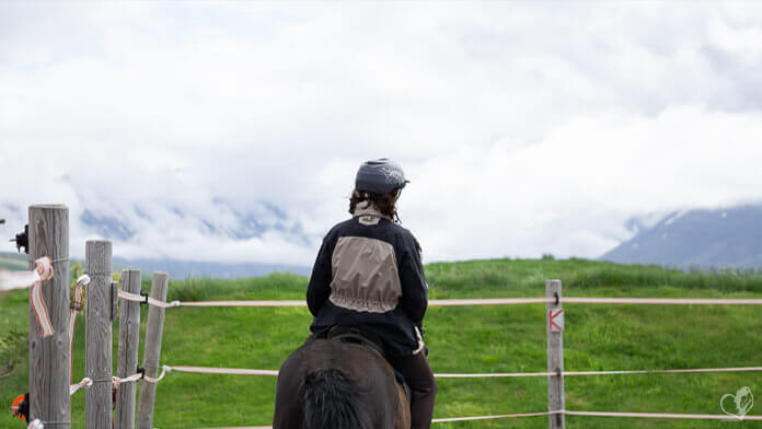 Eine Reiterin auf einem schwarzen Islandpferd. Sie reitet auf einem Platz inmitten grüner Wiesen, im Hintergrund sind wolkenverhangene Berge zu sehen.