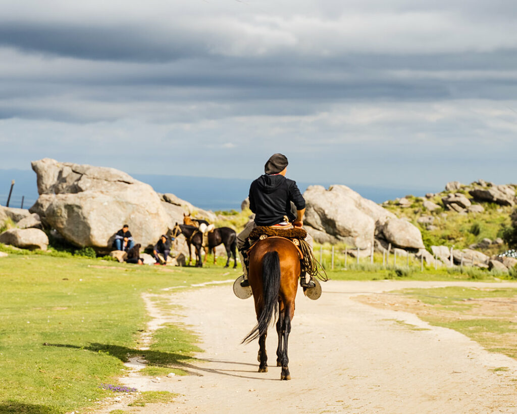 Ein junger Reiter auf dem Weg zu seinen Freunden, die man im Hintergrund an einem großen Felsen sitzen sieht, ihre Pferde neben ihnen.